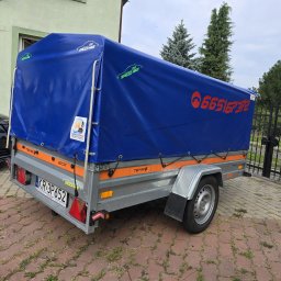 SPEED HADI TAXI MATEUSZ HADAŁA - Przyczepa samochodowa marki TEMA ECO z niebieską plandeką, na której widnieje numer telefonu oraz logo firmy transportowej 'Speed Hadi', zaparkowana na kostce brukowej przed ogrodzeniem.