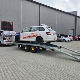 SPEED HADI TAXI MATEUSZ HADAŁA - Biały samochód taxi z napisem 'SPEED HADI' transportowany na lawecie z żółtymi felgami, zaparkowany na tle szarego budynku przemysłowego. W tle widoczny bus taxi z podobnym brandingiem.