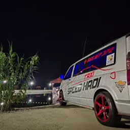 Srebrny bus taxi 'SPEED HADI' z czerwonymi felgami stoi nocą nad brzegiem oświetlonego stawu, z tyłu widoczny numer telefonu i logo 'TOW-TRUCK'.