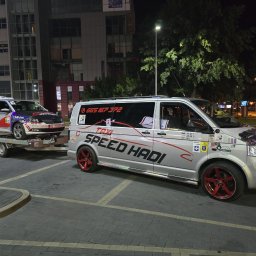 SPEED HADI TAXI MATEUSZ HADAŁA - Srebrny bus z czerwonymi felgami marki Volkswagen, oznaczony jako taksówka 'SPEED HADI', ciągnie na przyczepie osobowy samochód z taksometrem, również oznaczony jako taksówka, na tle nocnego...