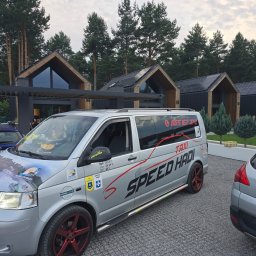 SPEED HADI TAXI MATEUSZ HADAŁA - Srebrny van z napisem 'TAXI SPEED HADI' i numerem telefonu, zaparkowany przed nowoczesnymi domami z ciemnego drewna i dużymi oknami, z naklejkami na masce i czerwonymi felgami.