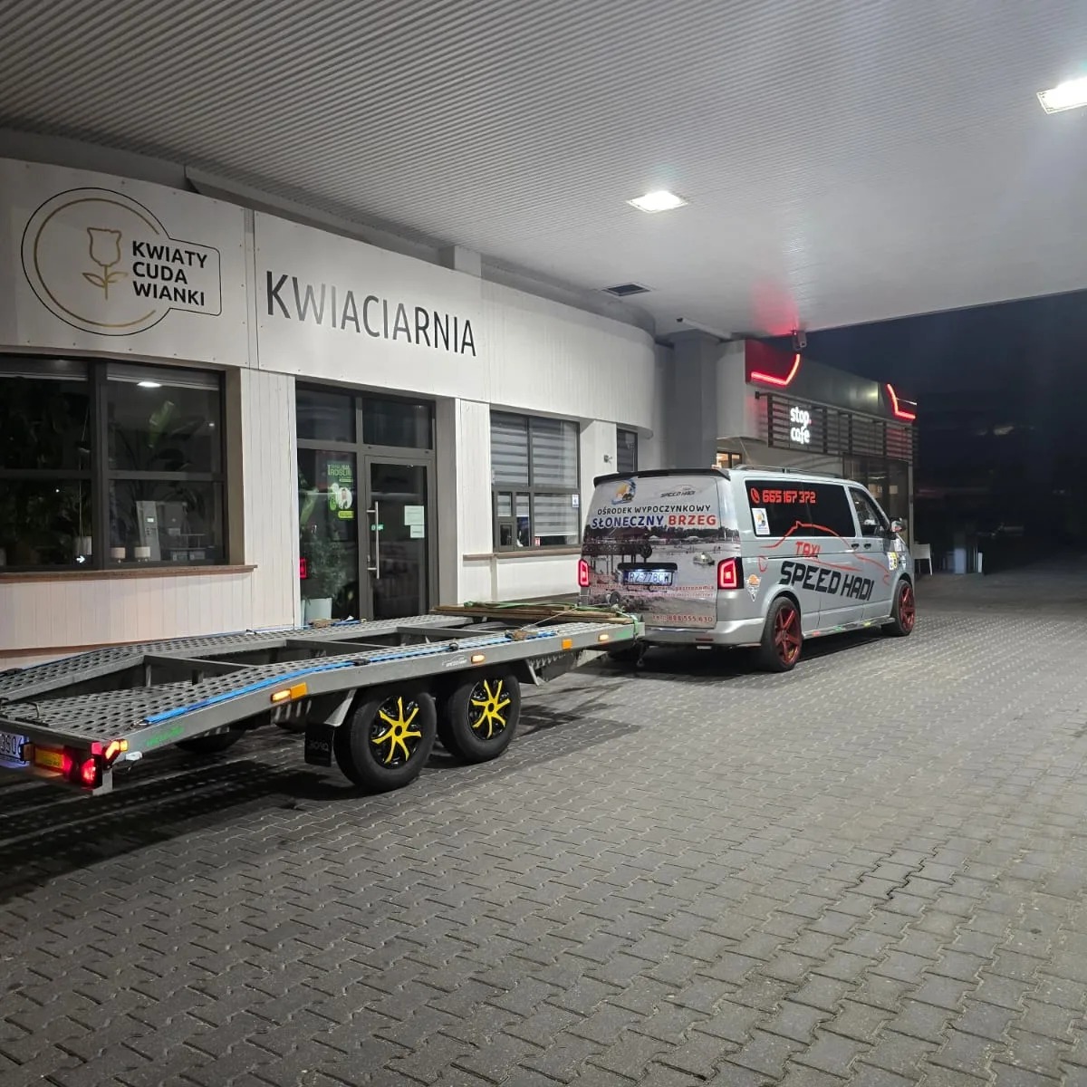 Laweta z żółtymi akcentami na kołach, zaparkowana przy stacji benzynowej w Rzeszowie, podpięta do srebrnego busa taxi z czerwonymi felgami i reklamą na karoserii.