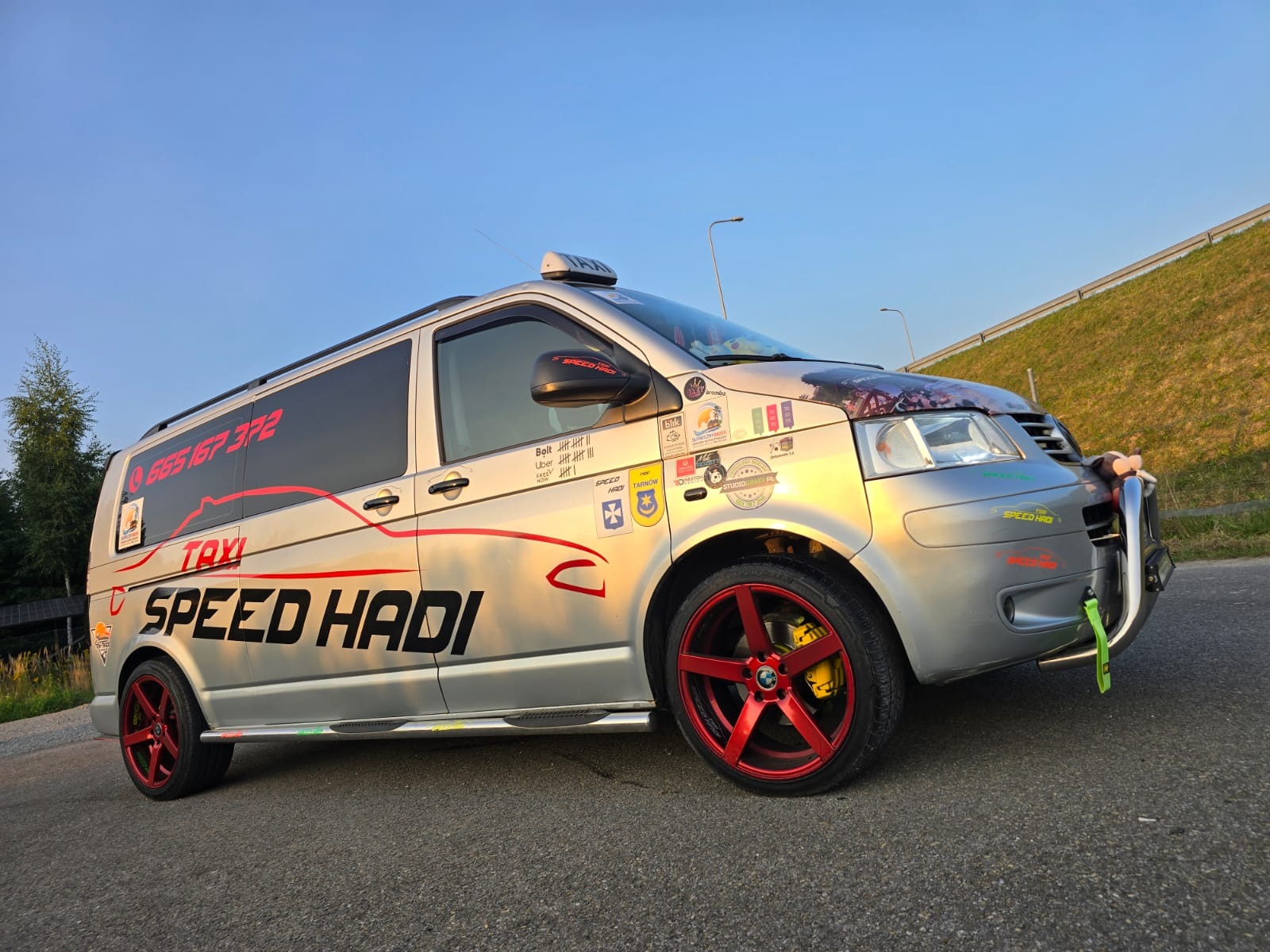 Srebrny bus z napisem 'TAXI SPEED HADI' i numerem telefonu, ozdobiony naklejkami i czerwonymi felgami, zaparkowany na asfalcie w słoneczny dzień.