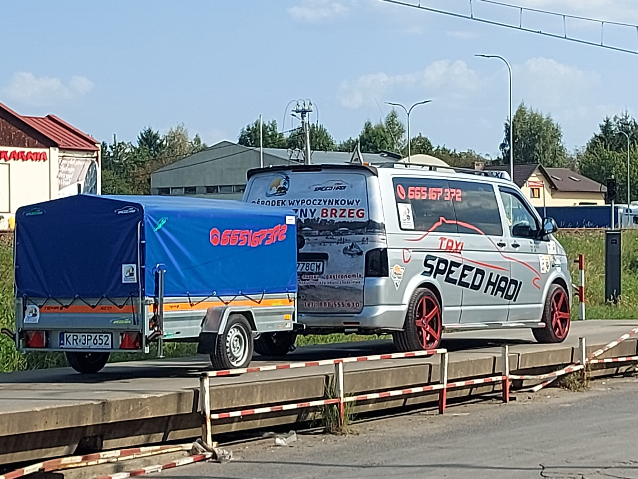 Srebrny bus z napisem 'TAXI SPEED HADI' ciągnie przyczepkę z niebieską plandeką, widoczne numery telefonu na busie i przyczepce, czerwone felgi w busie.