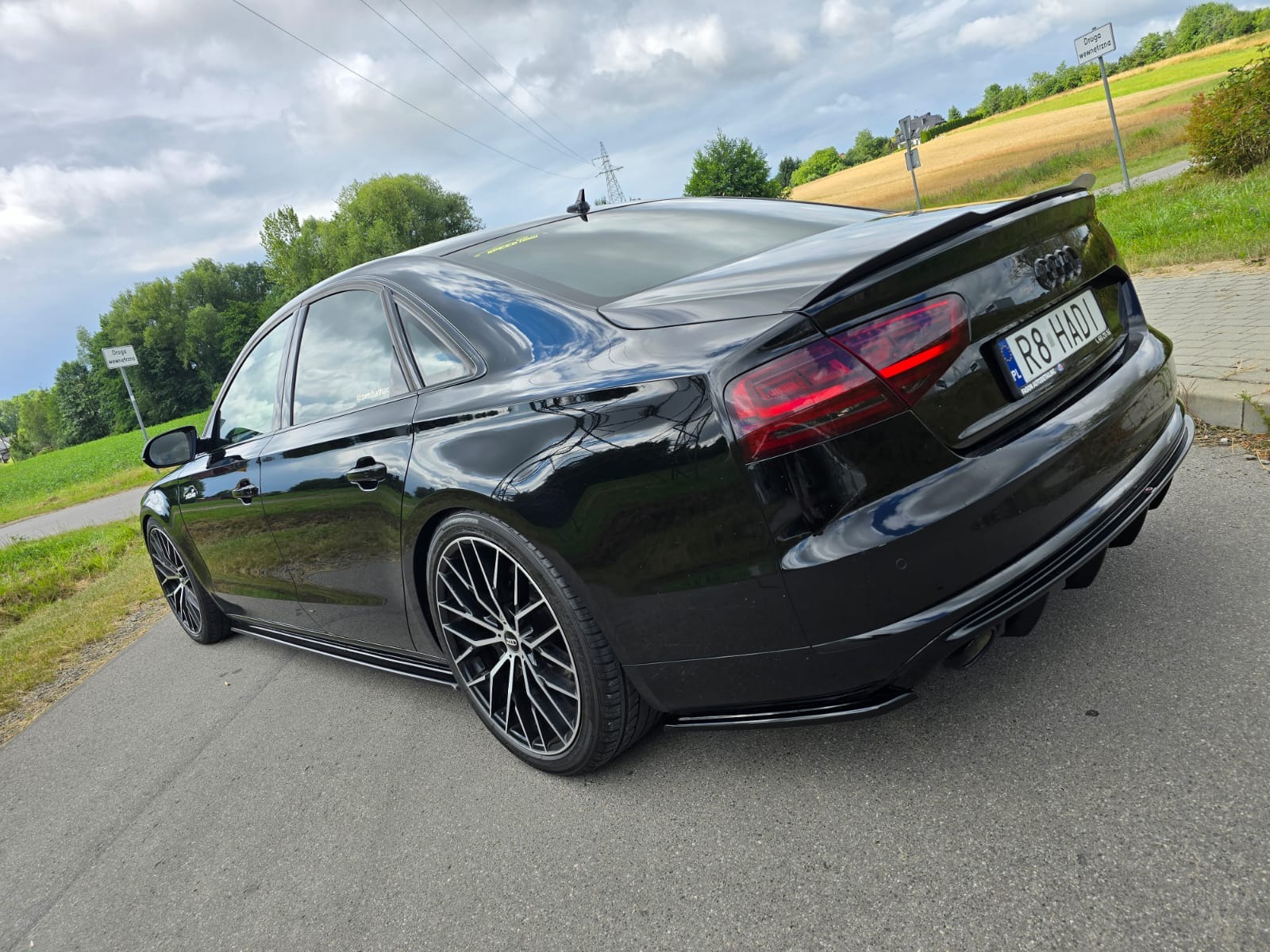 Czarny, zmodyfikowany samochód osobowy Audi A8 z polską tablicą rejestracyjną R8 HAD1, zaparkowany na asfaltowej drodze w wiejskim otoczeniu z polami i drzewami w tle.