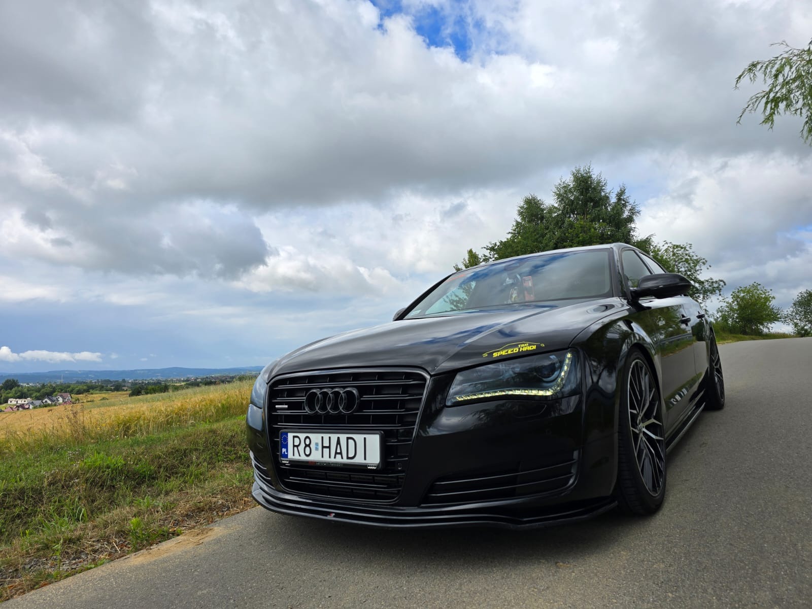 Czarna limuzyna Audi A8 z rejestracją R8 HADI, zaparkowana na poboczu drogi z widokiem na krajobraz, z naklejką 'SPEED HADI' na reflektorze.