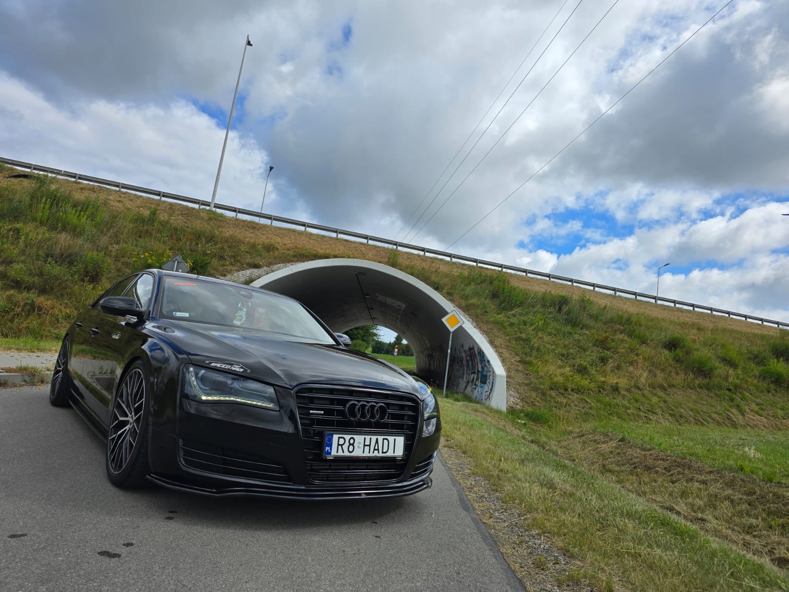 Czarna, luksusowa limuzyna Audi A8 z tuningowanymi felgami i naklejką na przedniej szybie, zaparkowana na drodze w pobliżu tunelu z graffiti i znakiem drogowym.