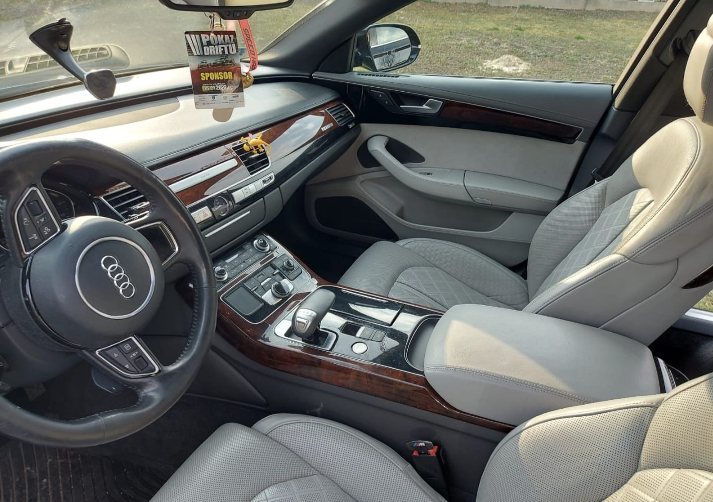 Wnętrze samochodu Audi A8 z jasną skórzaną tapicerką, drewnianymi wykończeniami i zawieszką 'Pokaz Driftu' na lusterku.