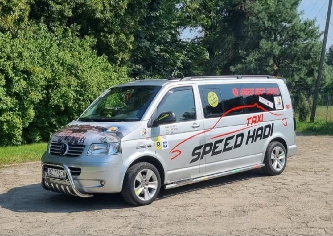 Srebrny bus z napisem 'SPEED HADI TAXI' i numerem telefonu, zaparkowany na żwirowym podłożu z zielonym tłem drzew.
