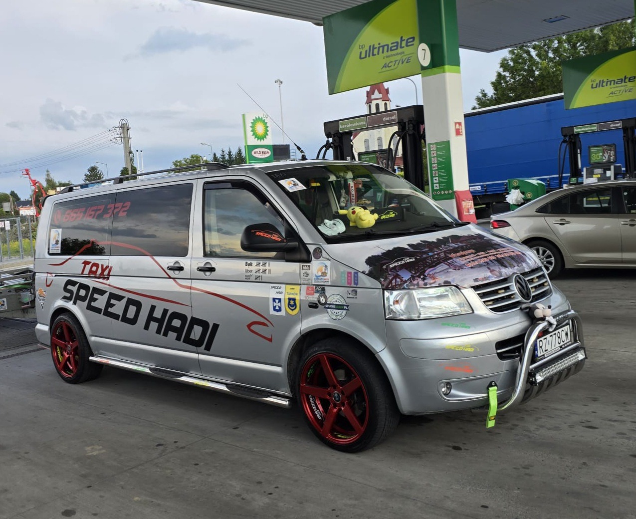 Srebrny van z czerwonymi felgami, oklejony naklejkami i grafikami, oznaczony jako TAXI 'SPEED HADI', zaparkowany przy dystrybutorze paliwa BP. Na desce rozdzielczej leży pluszowy Pikachu.