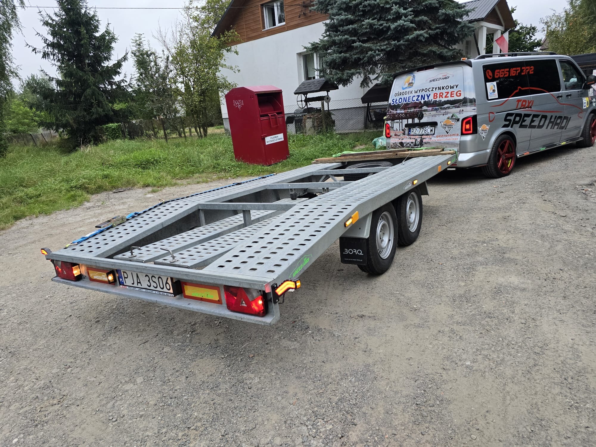 Szara laweta z metalową kratownicą i zapalonymi światłami, podłączona do srebrnego busa z reklamą ośrodka wypoczynkowego i numerem telefonu, stojąca na szutrowej drodze w wiejskiej scenerii...