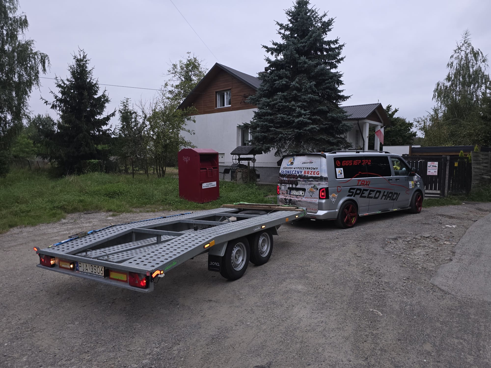 Szara laweta samochodowa marki BORO, podpięta do szarego busa z reklamami ośrodka wypoczynkowego 'Słoneczny Brzeg' i napisem 'TAXI SPEED HADI', stojąca na żwirowej drodze przed domem.