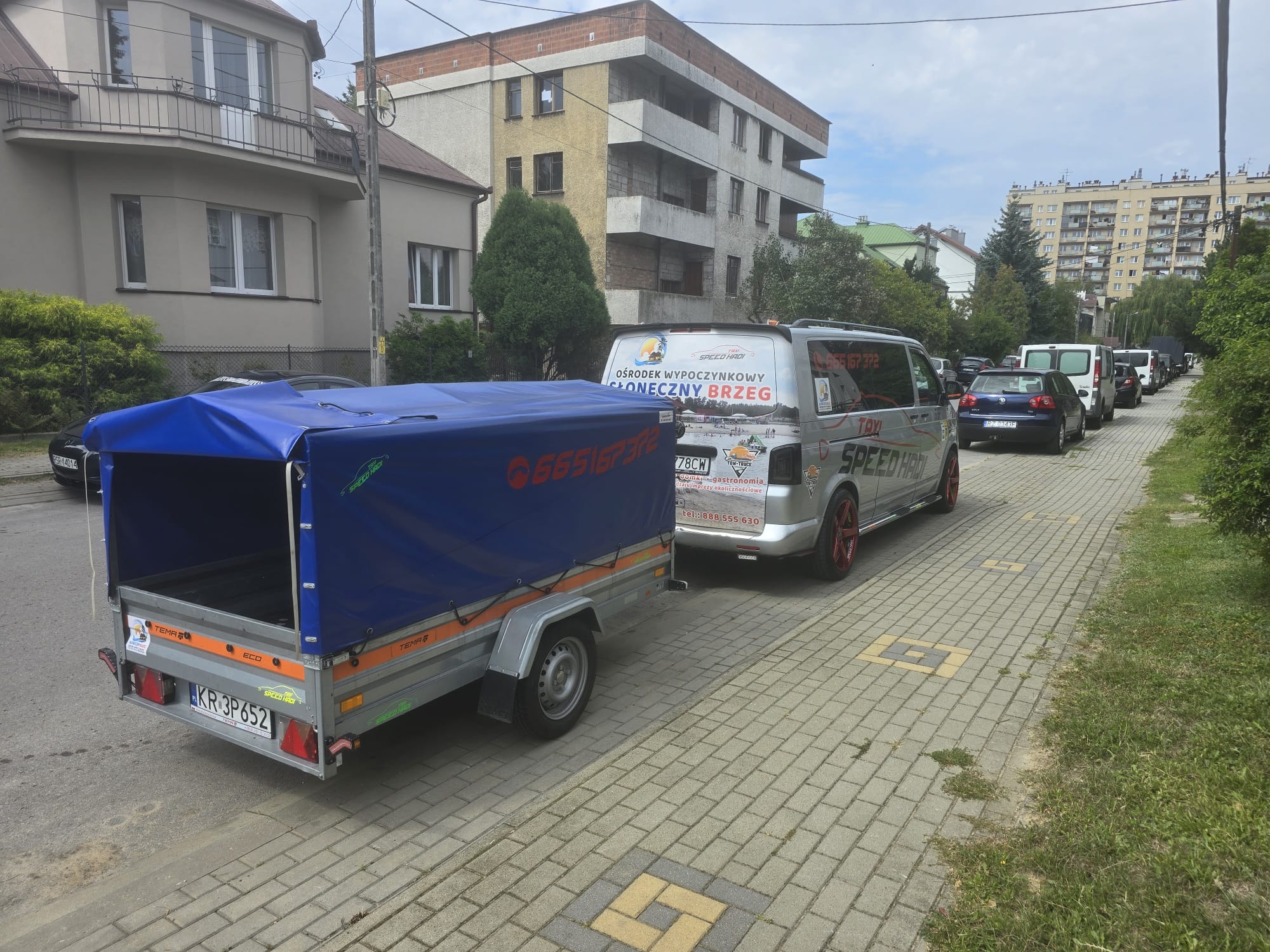 Przyczepa z niebieską plandeką zaparkowana na chodniku obok srebrnego busa z reklamą ośrodka wypoczynkowego i numerem telefonu.