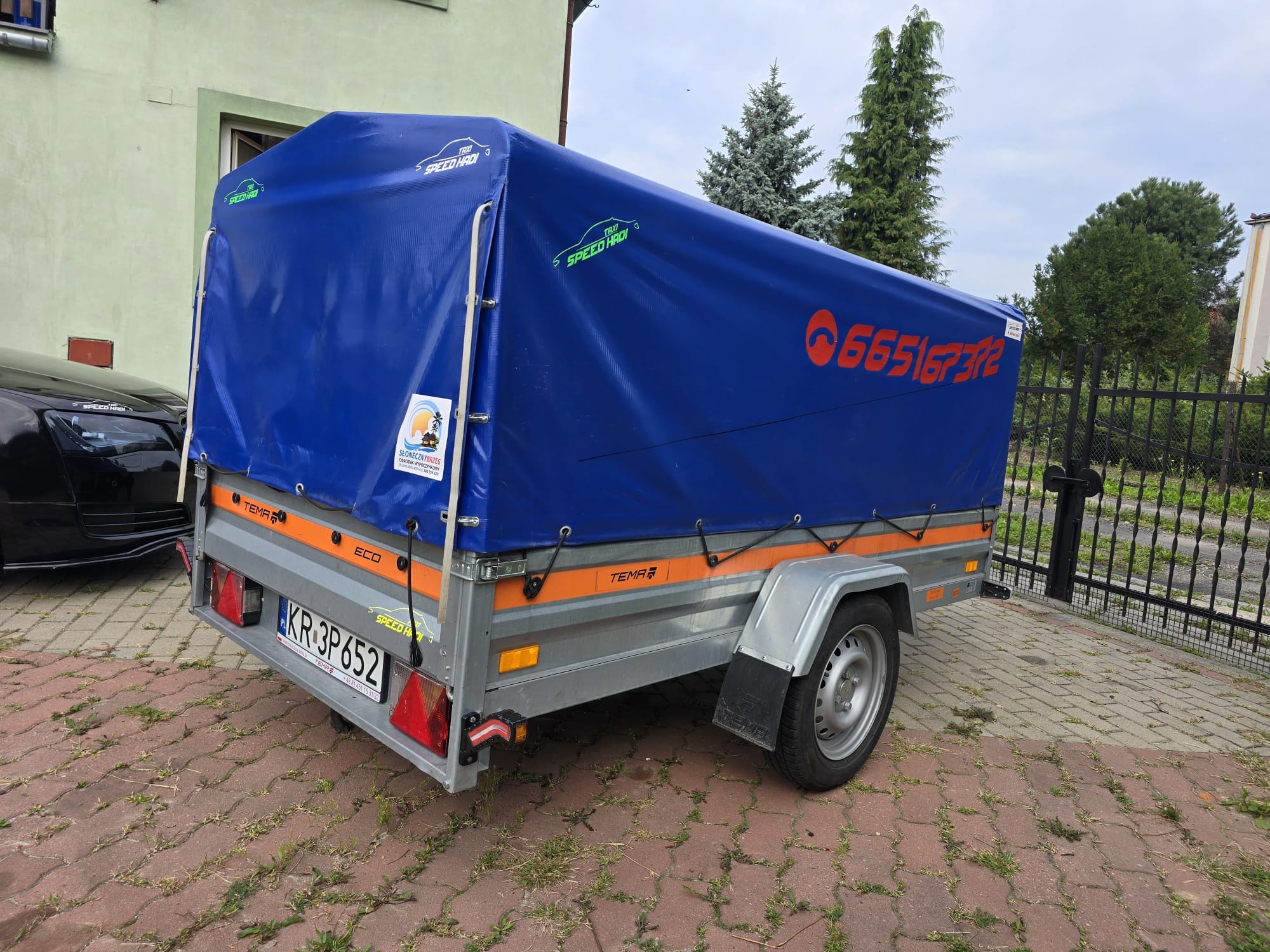 Przyczepa samochodowa marki TEMA ECO z niebieską plandeką, na której widnieje numer telefonu oraz logo firmy transportowej 'Speed Hadi', zaparkowana na kostce brukowej przed ogrodzeniem.