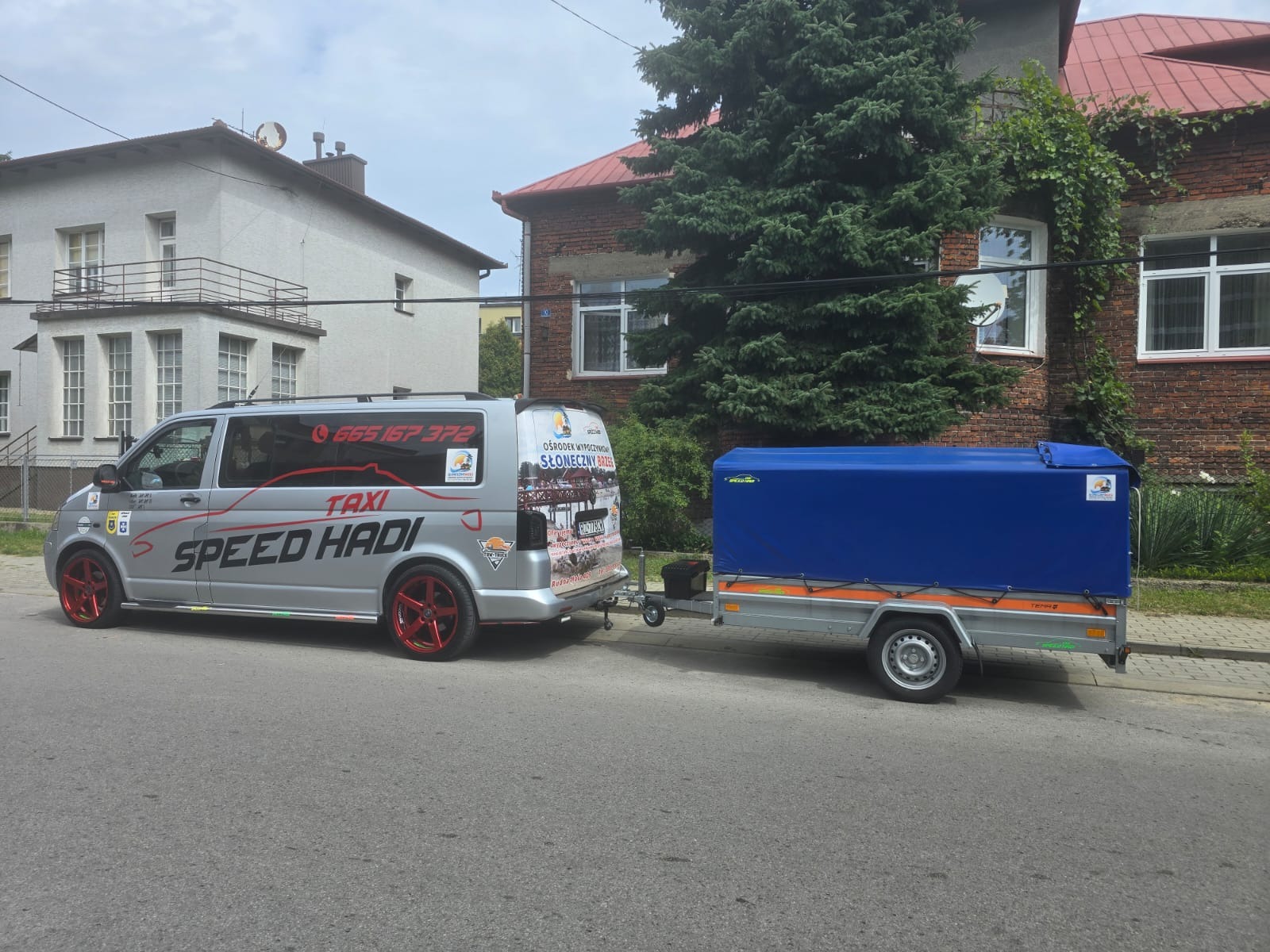 Srebrny bus taxi z czerwonymi felgami i przyczepką z niebieską plandeką zaparkowany na ulicy przed budynkami.