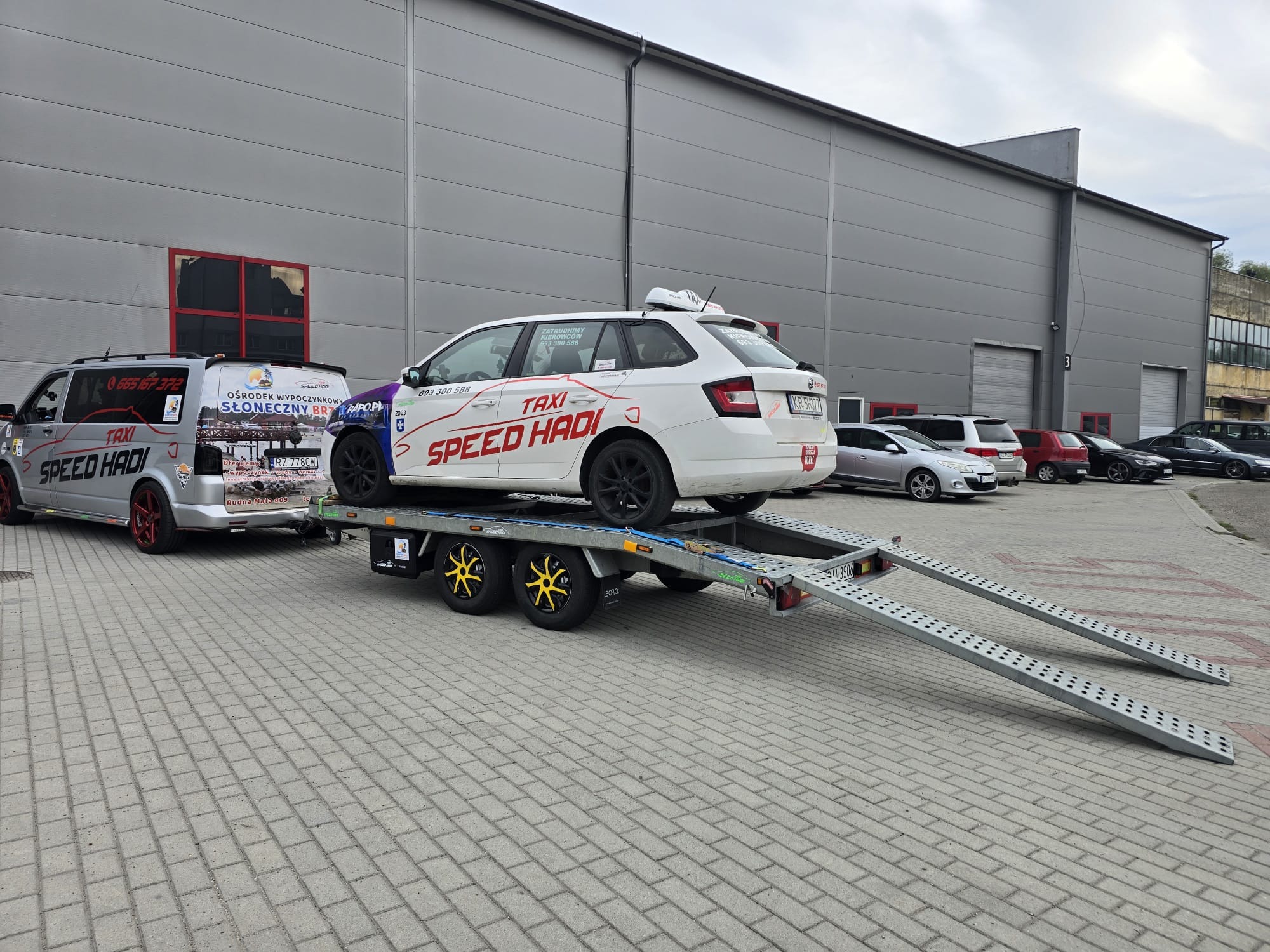 Biały samochód taxi z napisem 'SPEED HADI' transportowany na lawecie z żółtymi felgami, zaparkowany na tle szarego budynku przemysłowego. W tle widoczny bus taxi z podobnym brandingiem.