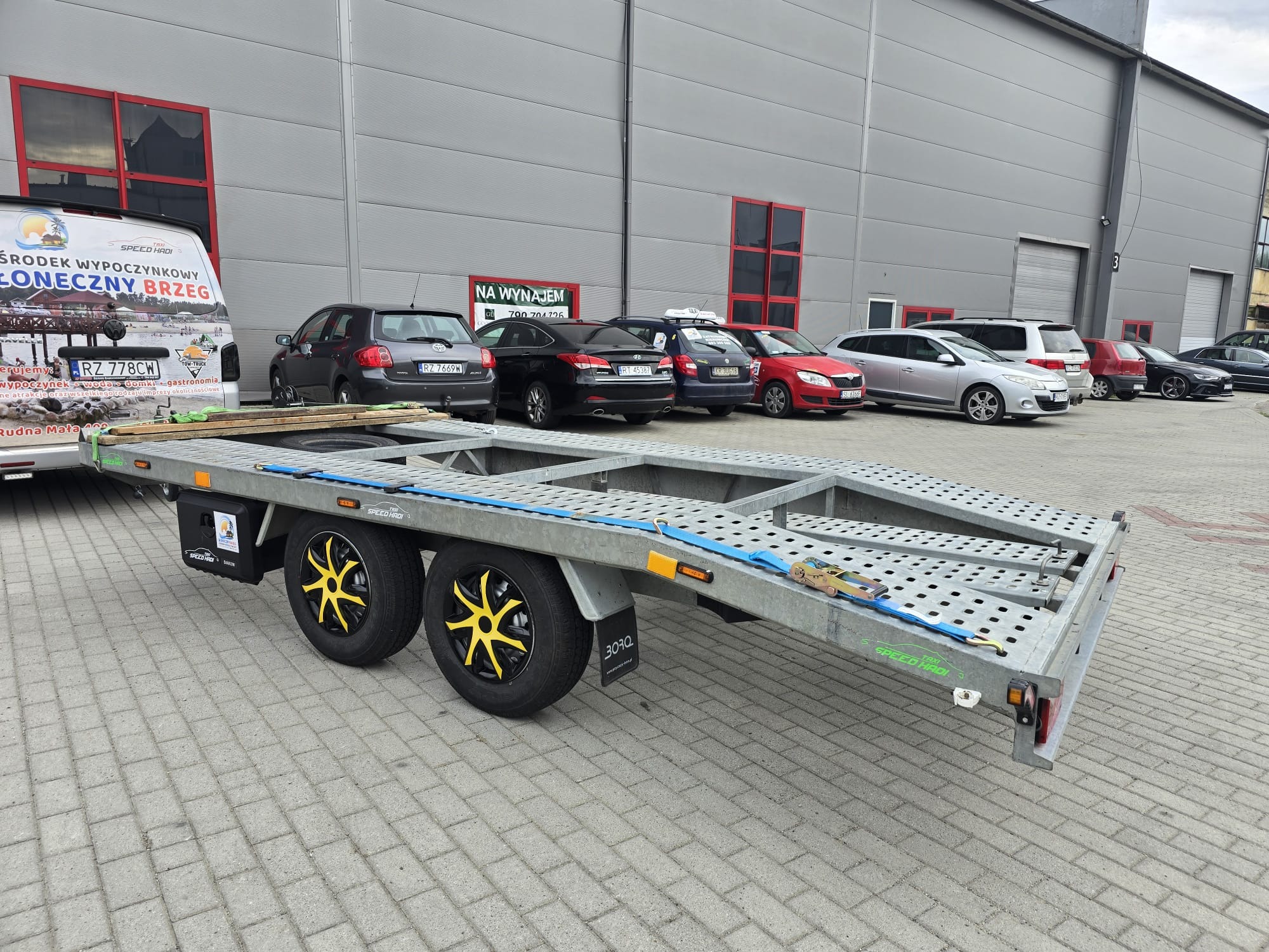 Pusta, dwuosiowa laweta samochodowa z żółtymi felgami w kształcie gwiazd, zaparkowana na kostce brukowej przed szarym budynkiem magazynowym, w tle zaparkowane samochody osobowe.