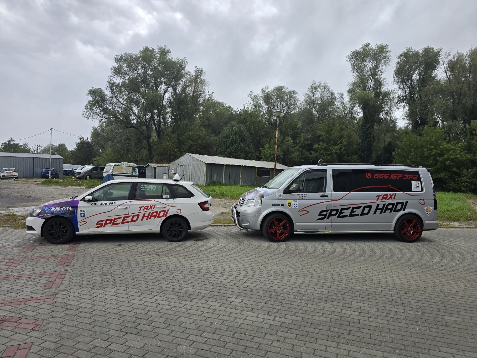 Dwa samochody taxi marki Skoda i Volkswagen, z logo 'Speed Hadi' i numerami telefonów, stojące obok siebie na parkingu z kostki brukowej, na tle zieleni i szarego nieba.