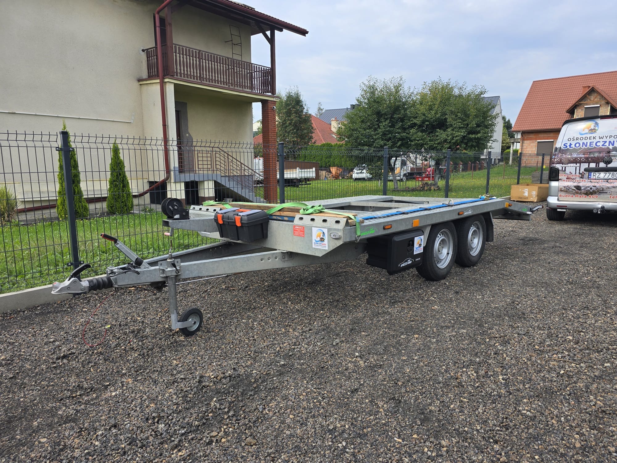 Pusta, dwuosiowa laweta z zamocowaną skrzynką narzędziową i pasami transportowymi, zaparkowana na żwirowym podłożu przed domem i częściowo zasłonięta przez busa z reklamą ośrodka wypoczynkowego.