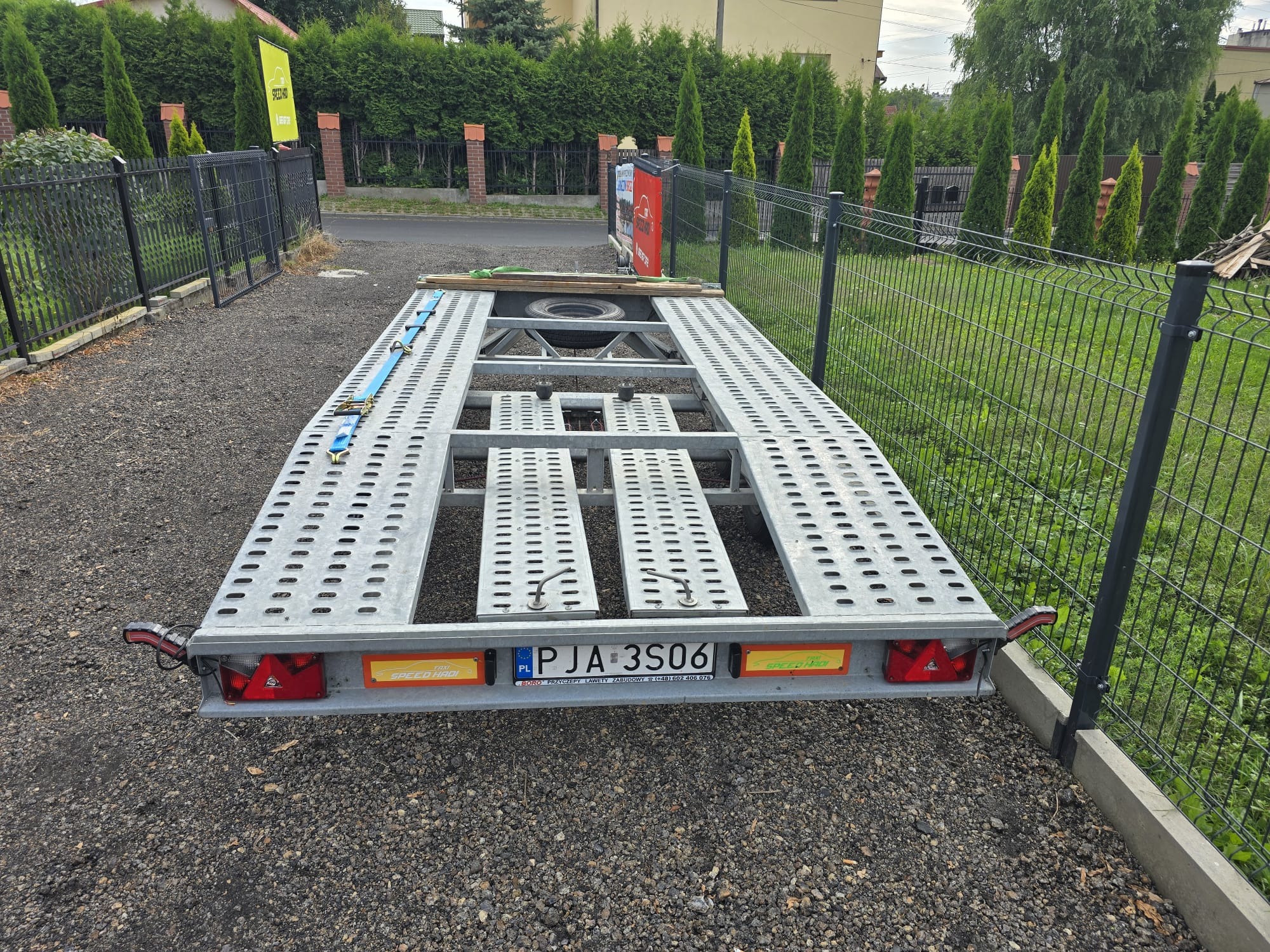 Pusta, srebrna laweta samochodowa zaparkowana na żwirowym podłożu, z widocznym kołem zapasowym, zabezpieczona niebieskim pasem transportowym, na tle ogrodzenia z siatki i zieleni.