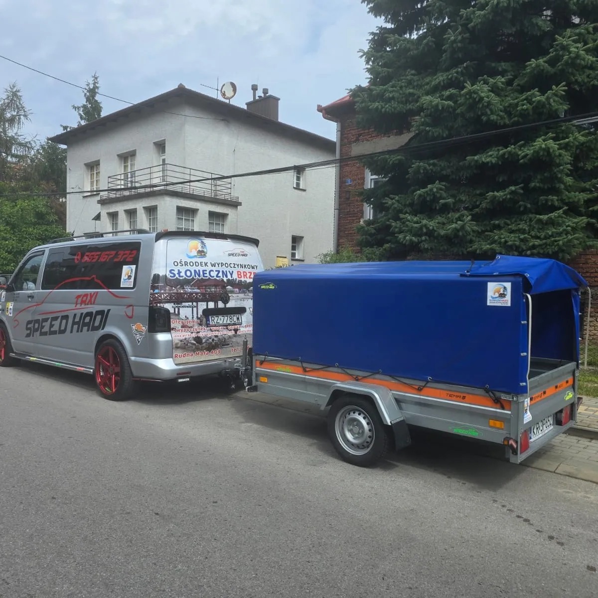 Srebrny bus z czerwonymi felgami i naklejkami reklamowymi, ciągnący niebieską przyczepę z plandeką, zaparkowany na ulicy w zabudowie miejskiej.