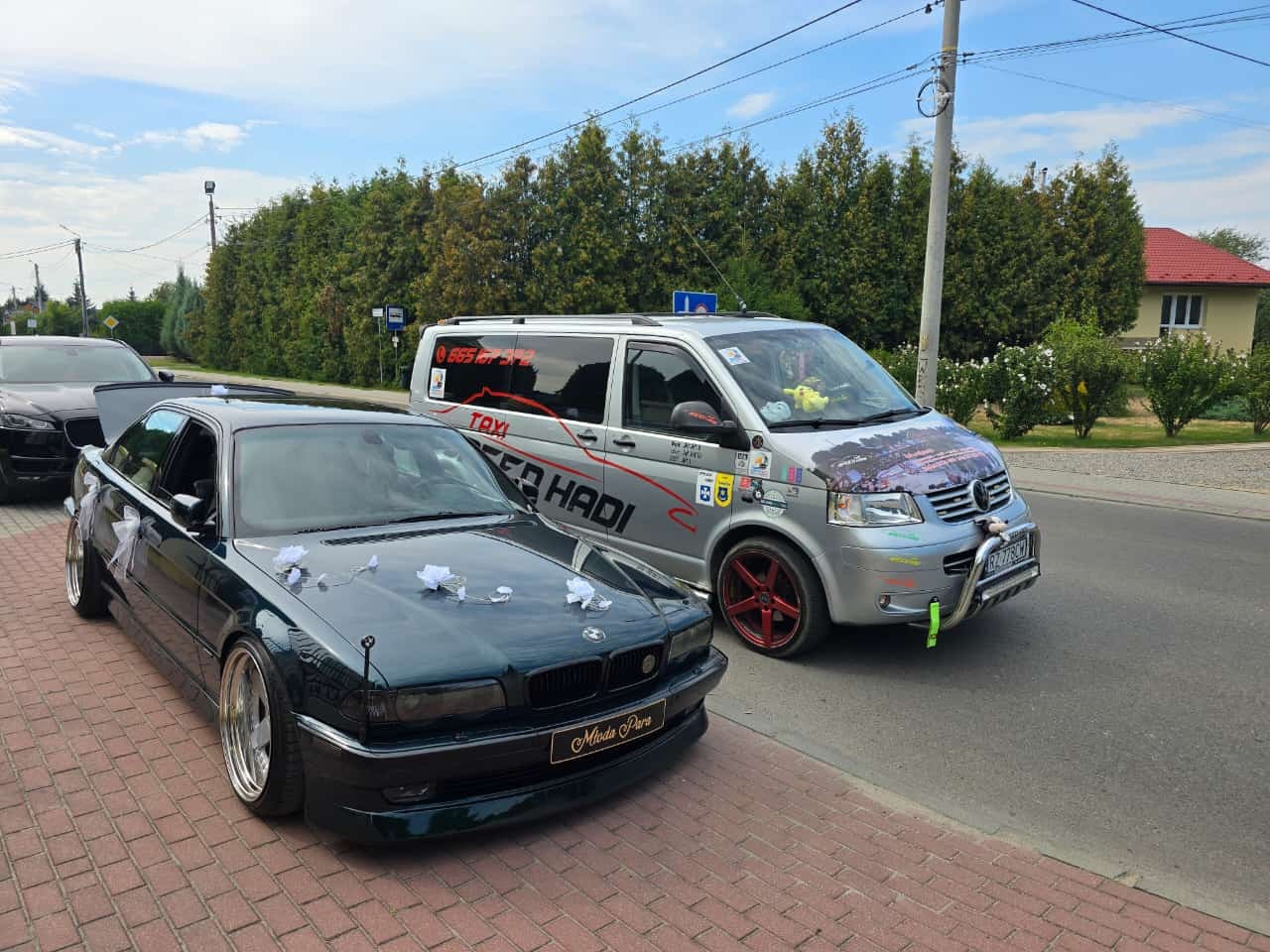Ciemnozielony samochód BMW udekorowany białymi kokardami ślubnymi i tabliczką 'Młoda Para' na tle srebrnego busa z napisem 'TAXI' i grafiką na masce, stojące na brukowanej drodze.