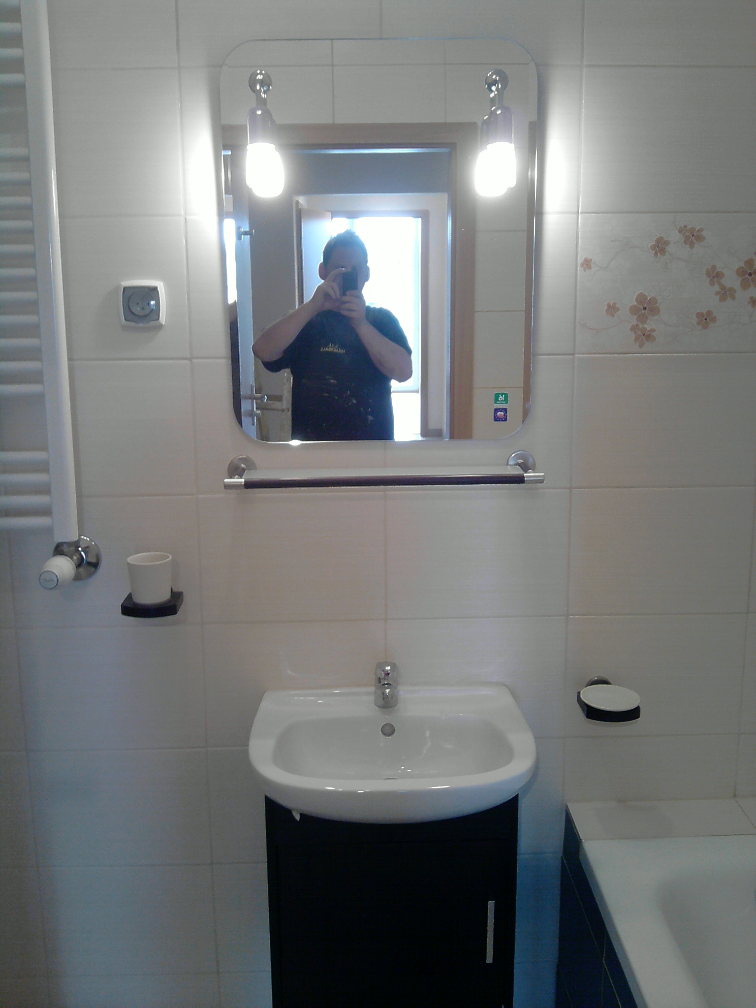 Umywalka z szafką w łazience z białymi płytkami i dekoracyjnym motywem kwiatowym, widoczny w lustrze fotografujący wnętrze mężczyzna.