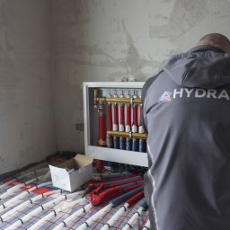 HYDRAKO Sławomir Kowal - Instalacja ogrzewania podłogowego w trakcie realizacji. Widoczne rury na folii grzewczej, rozdzielacz i instalator w bluzie z logo HydrakO.