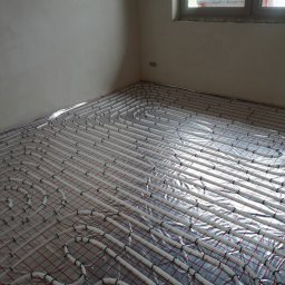 HYDRAKO Sławomir Kowal - Białe rury ogrzewania podłogowego, ułożone na folii z czerwoną siatką, w surowym wnętrzu z oknem. Widok z góry, ukośnie.