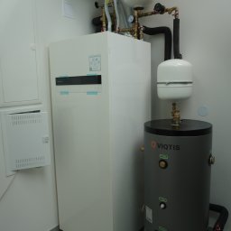 HYDRAKO Sławomir Kowal - Biała pompa ciepła Panasonic Aquarea i szary zasobnik Viqtis w kotłowni. Widoczne rury, zawory i osprzęt. Minimalistyczne wnętrze z szarą podłogą.