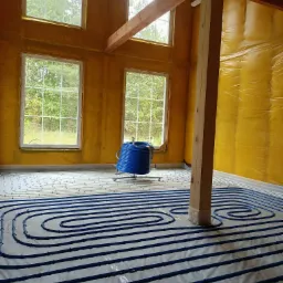 Wnętrze domu w trakcie instalacji ogrzewania podłogowego: widoczne niebieskie rury ułożone w pętle na podłożu, żółta folia na ścianach, drewniana konstrukcja stropu i kolumna. W tle okna z widokiem...