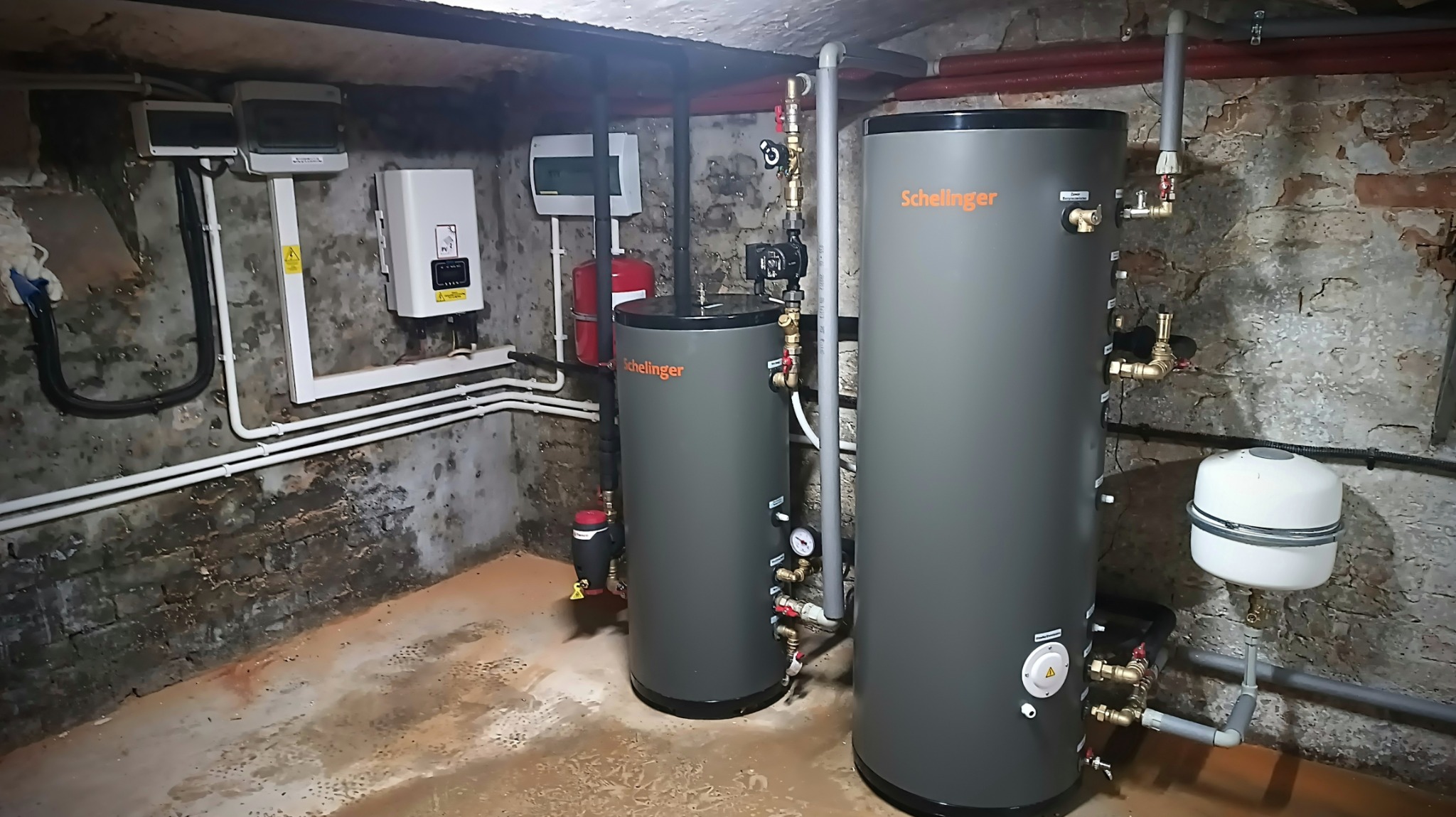 Dwa szare, cylindryczne zbiorniki Schelinger w piwnicy z odsłoniętą, starą cegłą, widoczne rury, zawory i manometry.