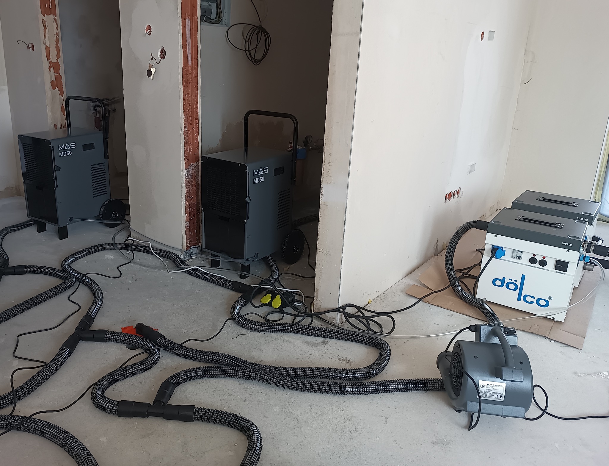 Osuszanie posadzek: dwa osuszacze MAS MD50 i dwa Dölco WVA-M z wentylatorem na surowym betonie, w tle ściany w trakcie wykańczania.