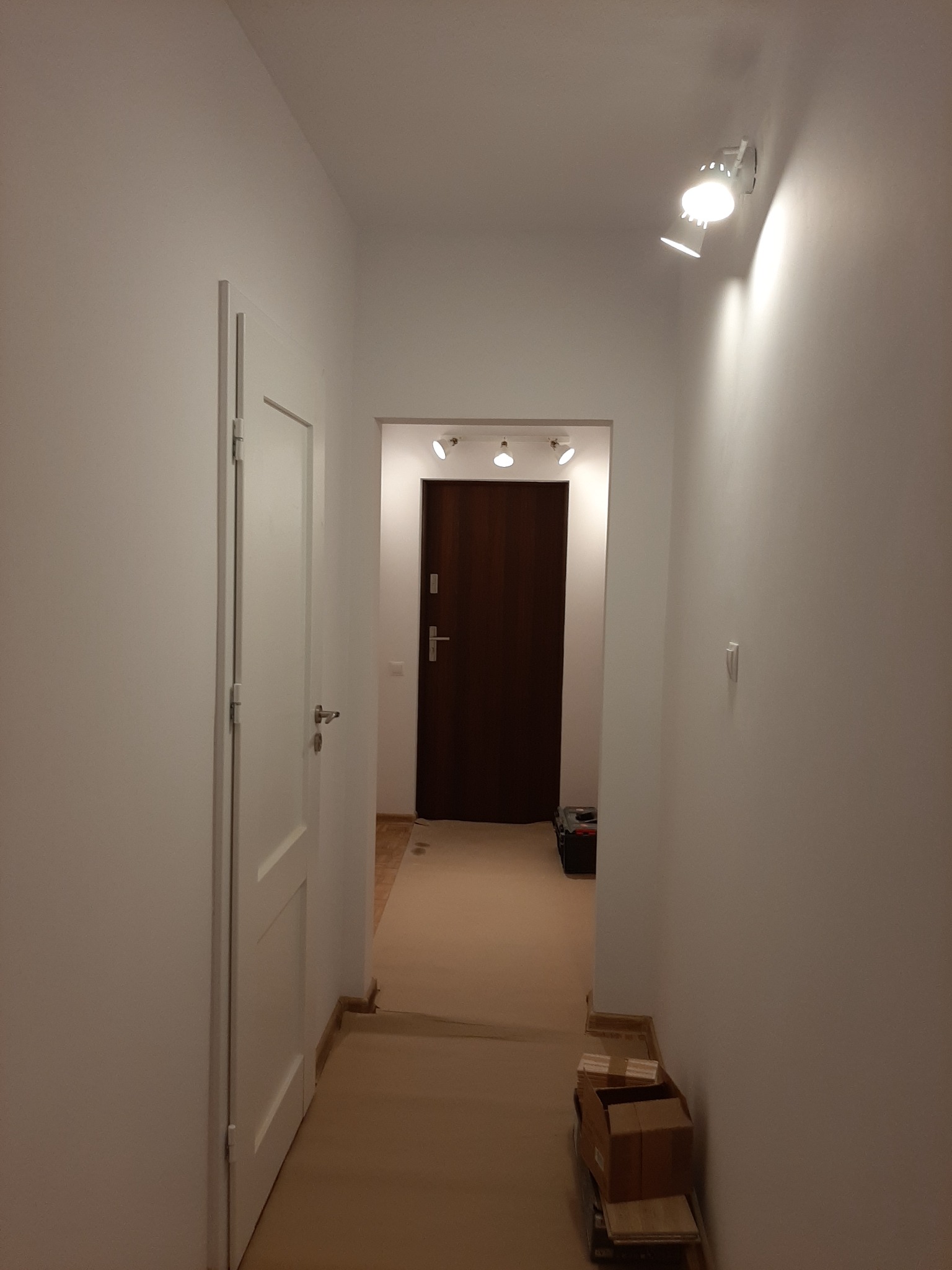 Wnętrze korytarza w trakcie remontu: świeżo pomalowane białe ściany, widoczna skrzynka narzędziowa w głębi, kartony z materiałami budowlanymi na podłodze zabezpieczonej kartonem, białe drzwi po...