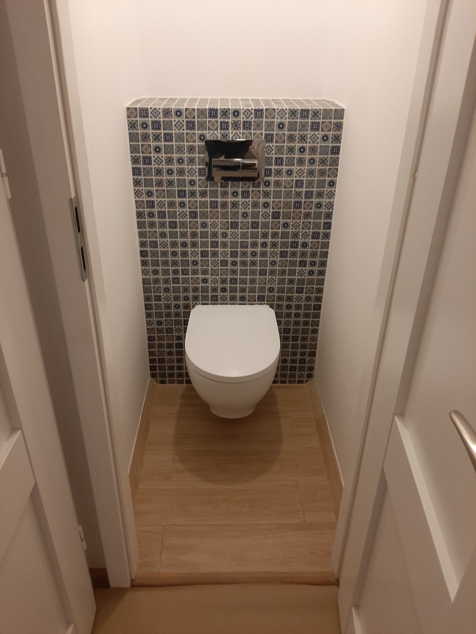 Nowoczesna toaleta podwieszana z geometryczną mozaiką nad spłuczką, drewnopodobna podłoga i białe ściany, widok zza otwartych drzwi.