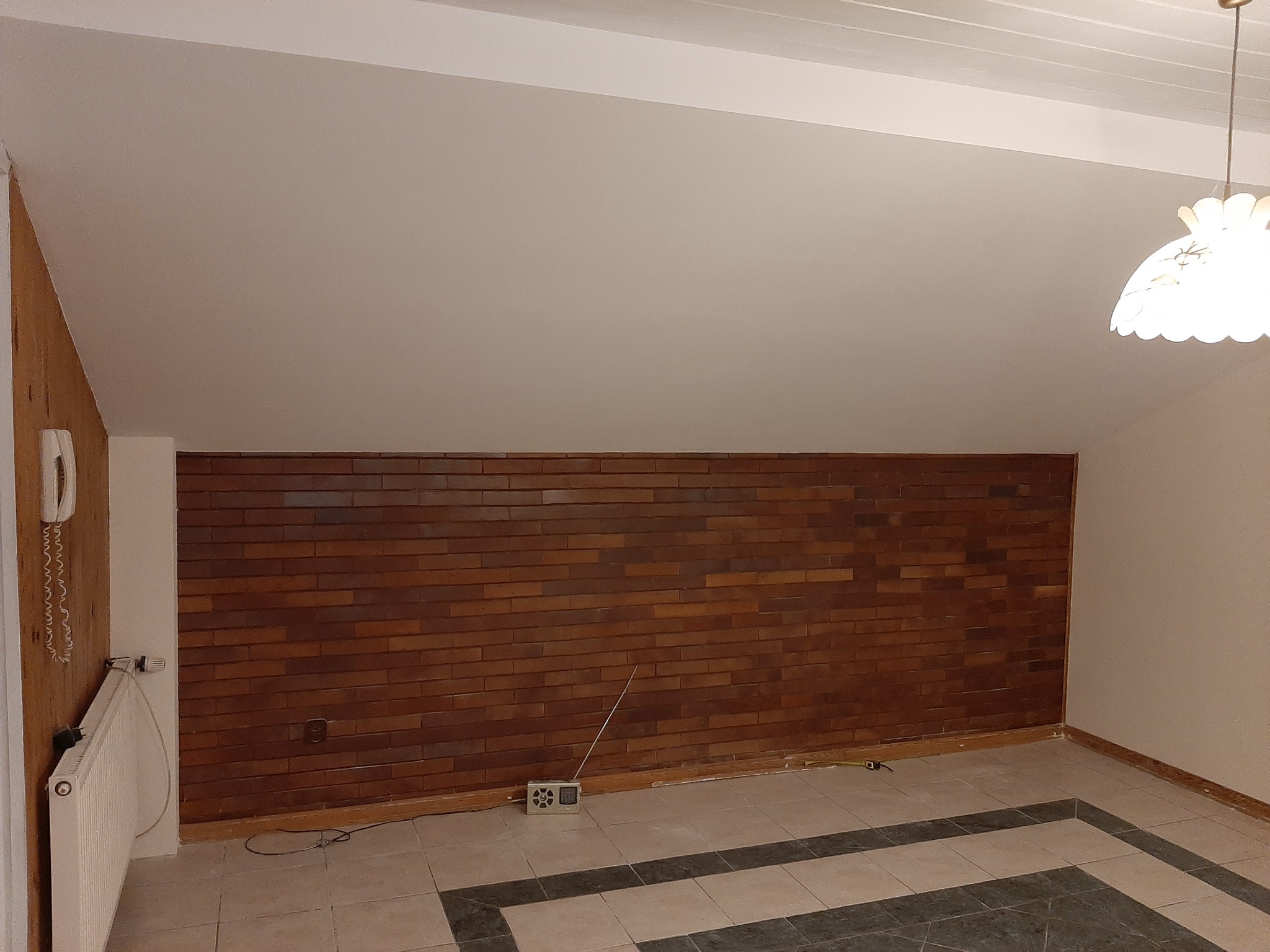 Pokój na poddaszu z brązową ścianą z płytek imitujących cegłę, białą skośną ścianą, grzejnikiem i starą radiem na podłodze z płytek.