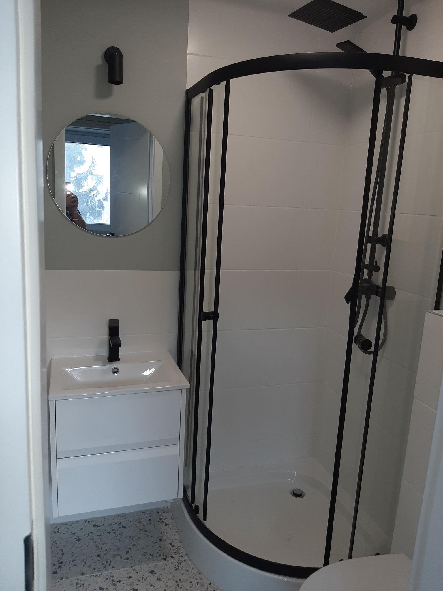 Nowoczesna łazienka po remoncie w Warszawie: białe płytki, kabina prysznicowa z czarnymi profilami, okrągłe lustro nad białą szafką z umywalką i czarną armaturą.