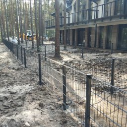 Artcast - Częściowo zamontowane, czarne ogrodzenie panelowe na piaszczystym terenie z budynkiem w tle otoczonym drzewami. Widoczny sprzęt budowlany.