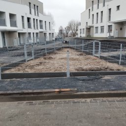 Artcast - Nowoczesne osiedle z szarą siatką ogrodzeniową, świeżo zamontowaną na tle minimalistycznej architektury. Widoczne przygotowane podłoże i kostka brukowa.