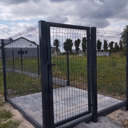 Artcast - Otwarta furtka z panelowego ogrodzenia w kolorze antracytowym, z widocznym podłożem z kostki brukowej i domem w tle, w pochmurny dzień.