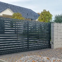 Nowoczesna, przesuwna brama wjazdowa z poziomych, szarych paneli, osadzona w słupkach z szarej kostki. Tło: dom z ciemnym dachem i jesienne drzewa.