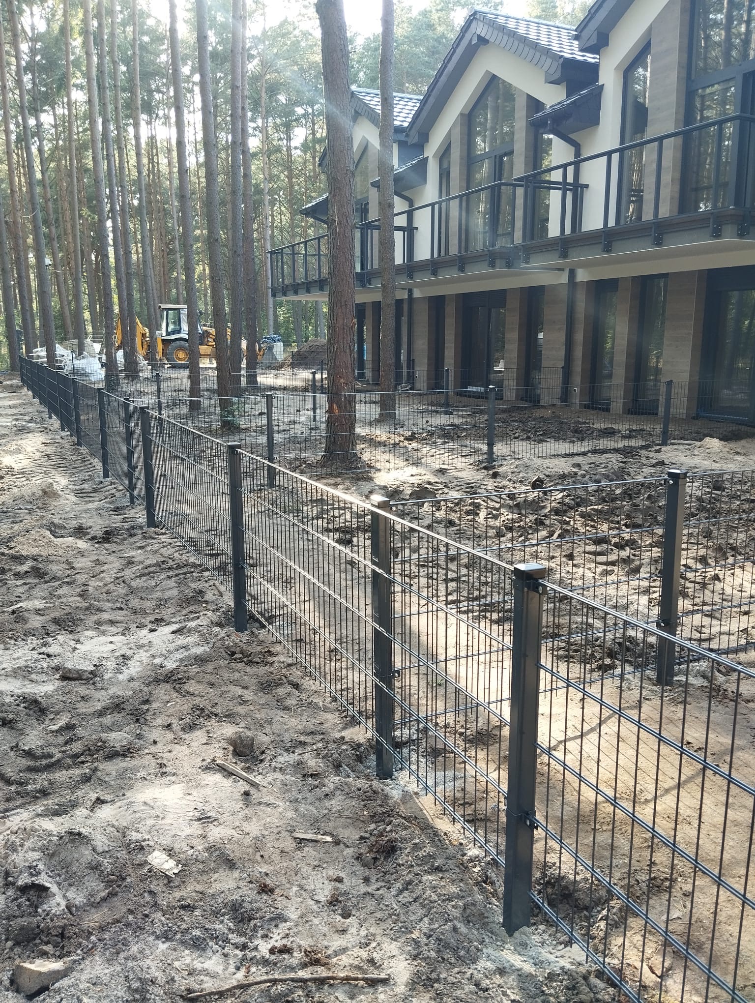 Częściowo zamontowane, czarne ogrodzenie panelowe na piaszczystym terenie z budynkiem w tle otoczonym drzewami. Widoczny sprzęt budowlany.