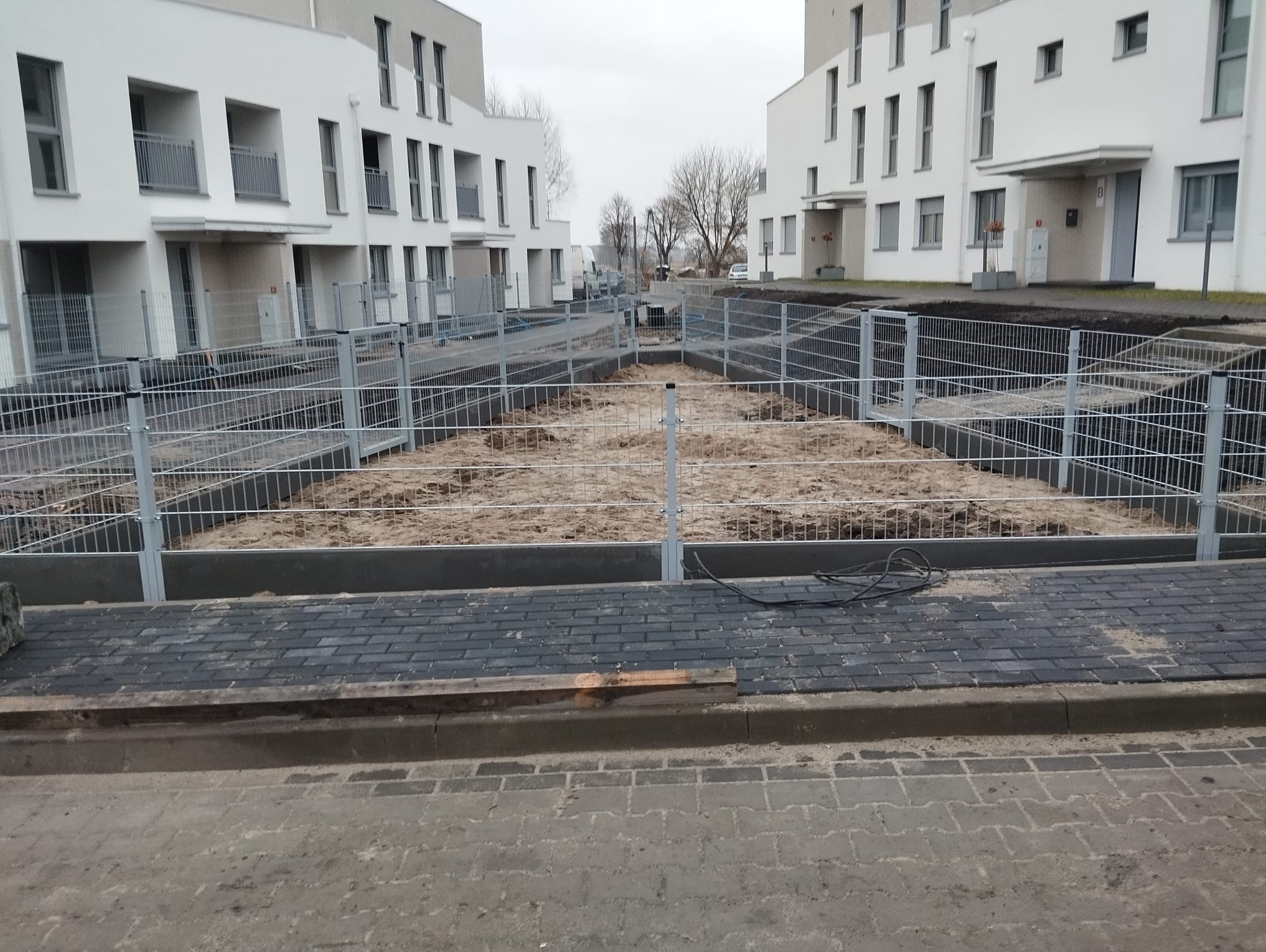 Nowoczesne osiedle z szarą siatką ogrodzeniową, świeżo zamontowaną na tle minimalistycznej architektury. Widoczne przygotowane podłoże i kostka brukowa.