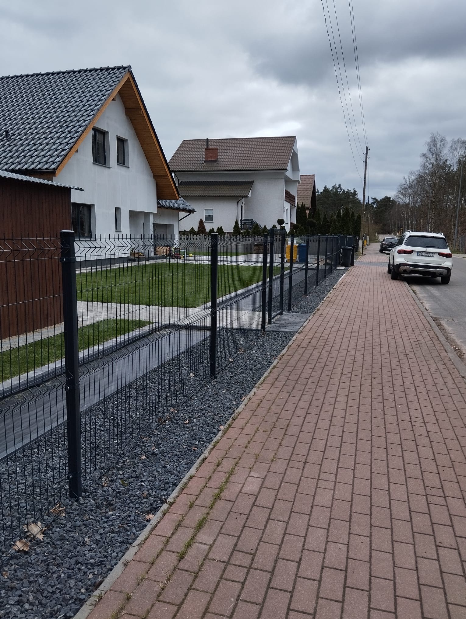Nowoczesne ogrodzenie panelowe w kolorze antracytowym, oddzielające posesję od chodnika z kostki. W tle widoczne domy jednorodzinne i zaparkowany samochód.
