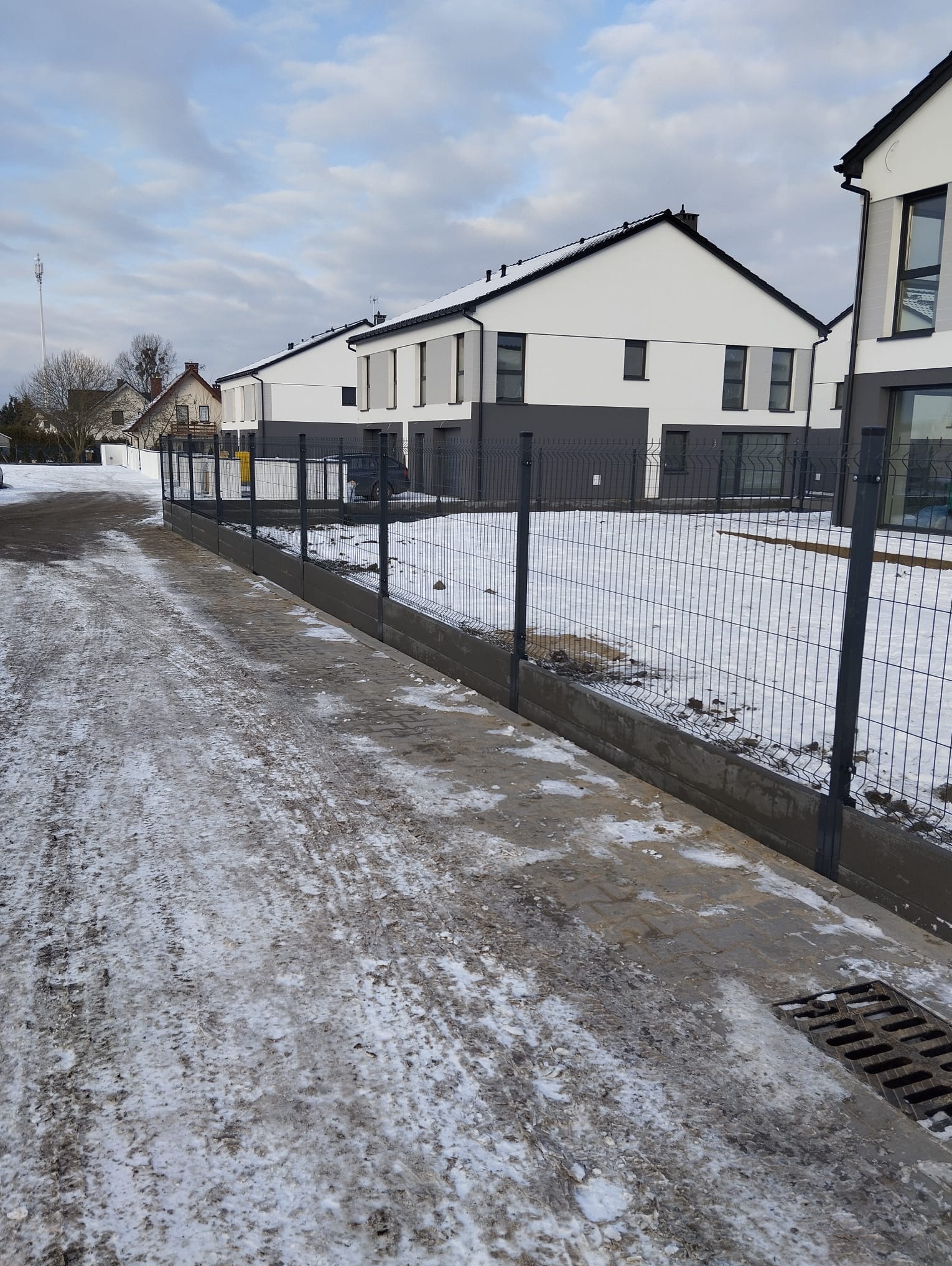 Nowoczesne ogrodzenie panelowe w kolorze grafitowym, zamontowane na betonowej podmurówce, oddziela posesję od ośnieżonej drogi. W tle widoczne domy.