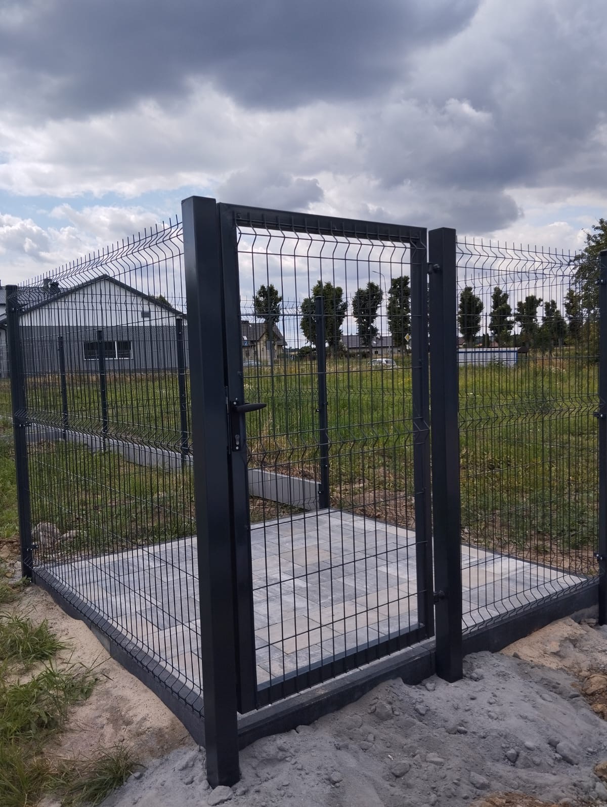 Otwarta furtka z panelowego ogrodzenia w kolorze antracytowym, z widocznym podłożem z kostki brukowej i domem w tle, w pochmurny dzień.