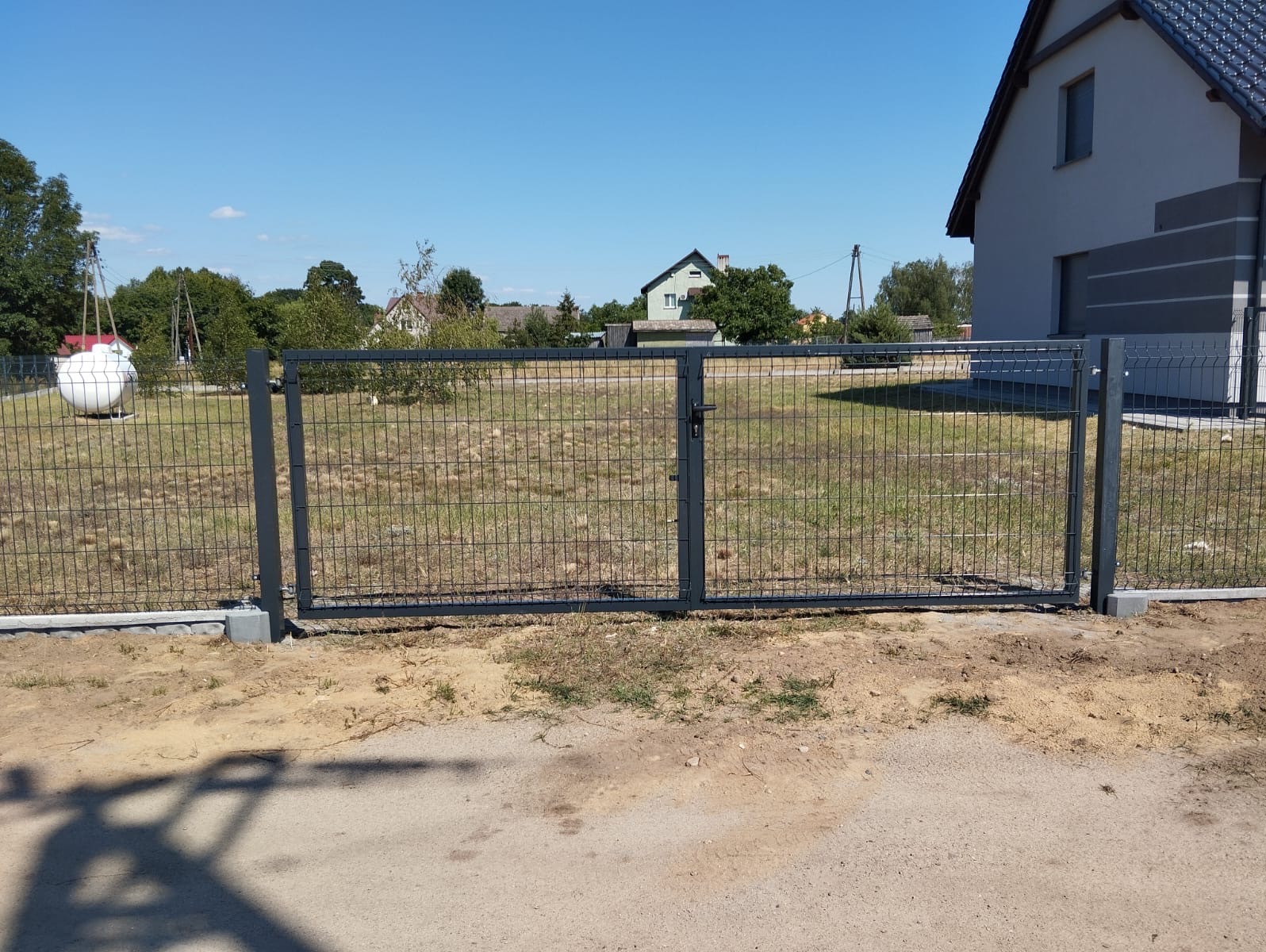 Nowoczesna, grafitowa brama panelowa z siatki, zamykająca posesję. W tle dom z szarą elewacją i zielone drzewa.
