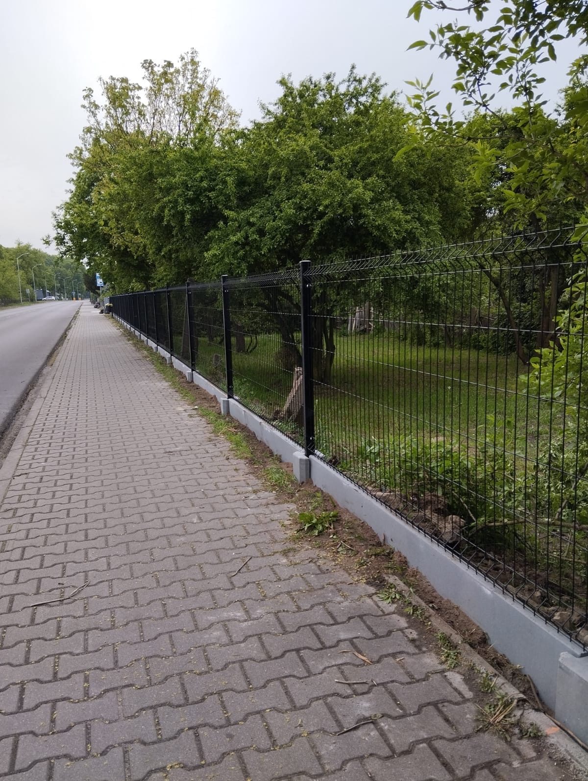 Długi, czarny panel ogrodzeniowy z betonową podmurówką, ciągnący się wzdłuż chodnika z kostki brukowej, za nim zieleń drzew i krzewów. Perspektywa z boku.