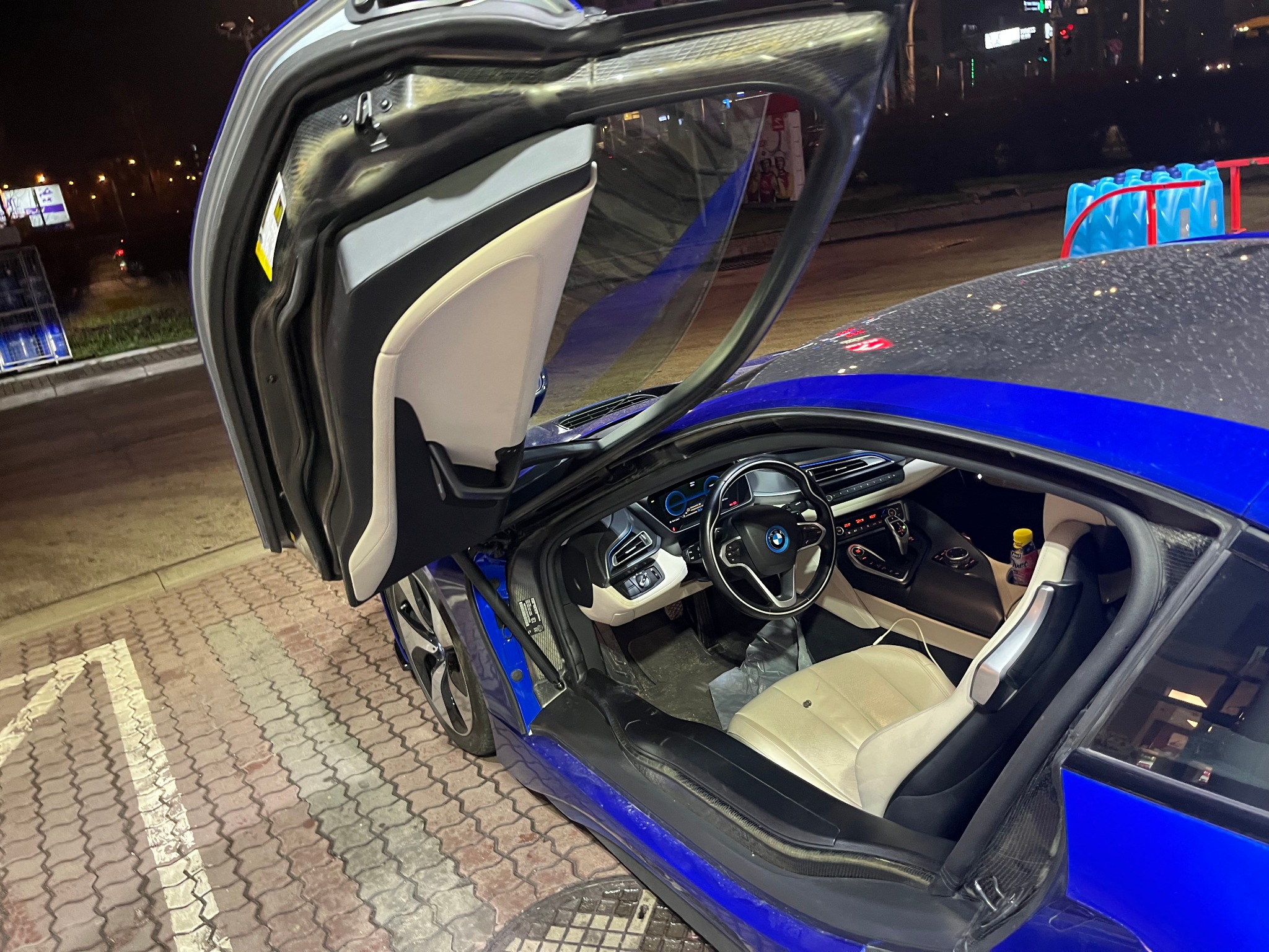 Nocne ujęcie otwartych drzwi motylkowych niebieskiego BMW i8, widoczne wnętrze z jasną tapicerką, na zewnątrz oświetlona ulica.