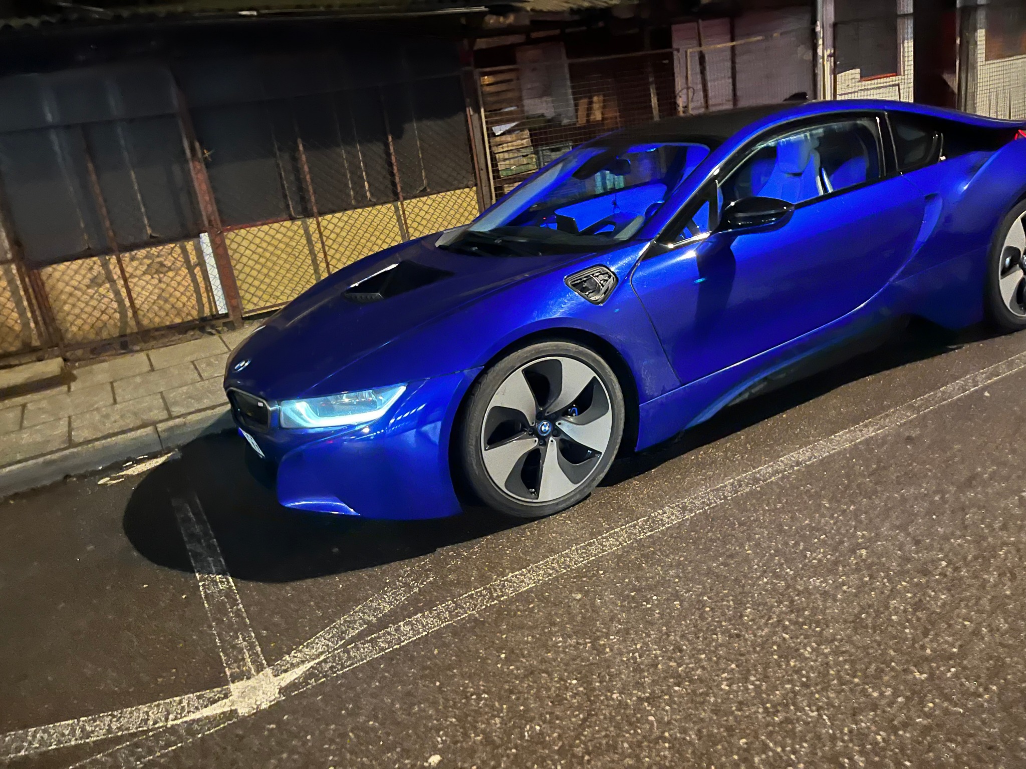 Błyszczący, granatowy samochód sportowy BMW i8 zaparkowany na ulicy z widocznymi znakami poziomymi, sfotografowany nocą.